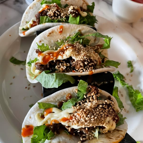 Four Vietnamese tacos with beef, lettuce, sesame seeds, and sauces on a white plate.